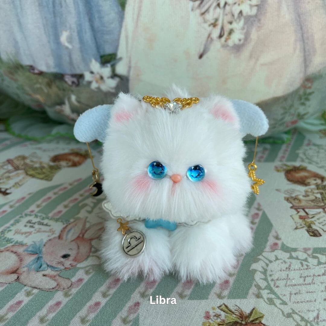 Libra kitten plush toy with blue eyes, white faux fur, celestial accessories, and zodiac-themed charm on a decorative background.
